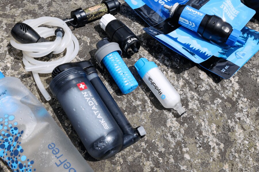 Sada různých přenosných vodních filtrů Sawyer, HydraPak a Katadyn na kamenném podkladu. Foto: Rigad