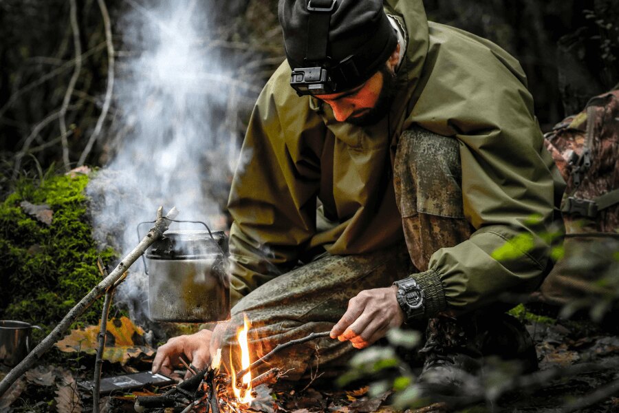 Muž v membránové bundě u ohně v lese. Zdroj: https://www.pexels.com/photo/kneeling-man-starting-fire-in-a-fireplace-15538110/
