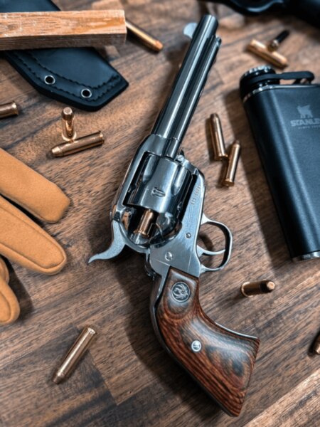 Jednočinný revolver Ruger s dlouhou hlavní a dřevěnou rukojetí položený mezi náboji a outdoorovým vybavením.