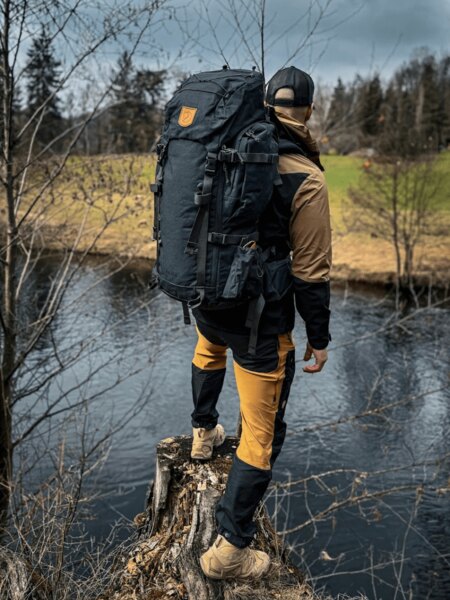 Trekař s plně naloženým batohem stojí u řeky na okraji lesa, připravený na vícedenní pochod. Foto: Rigad