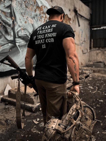 Muž v tričku The Trace s motivem CQB drží pušku a taktický batoh v industriálním prostředí.