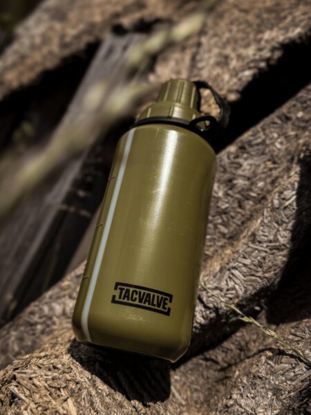 Polní láhev TacValve® ModCan v zeleném provedení položená na dřevěném podkladu v outdoorovém prostředí.