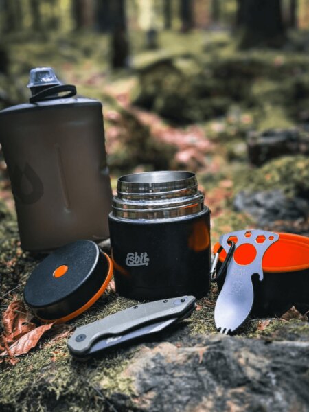 Otevřená jídelní termoska Esbit s outdoorovou lahví, skládacím nožem a příborem v lese.