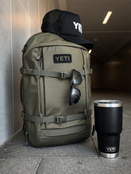 Světle šedý batoh YETI Ranchero 27 l s čepicí a černou lahví YETI Rambler.