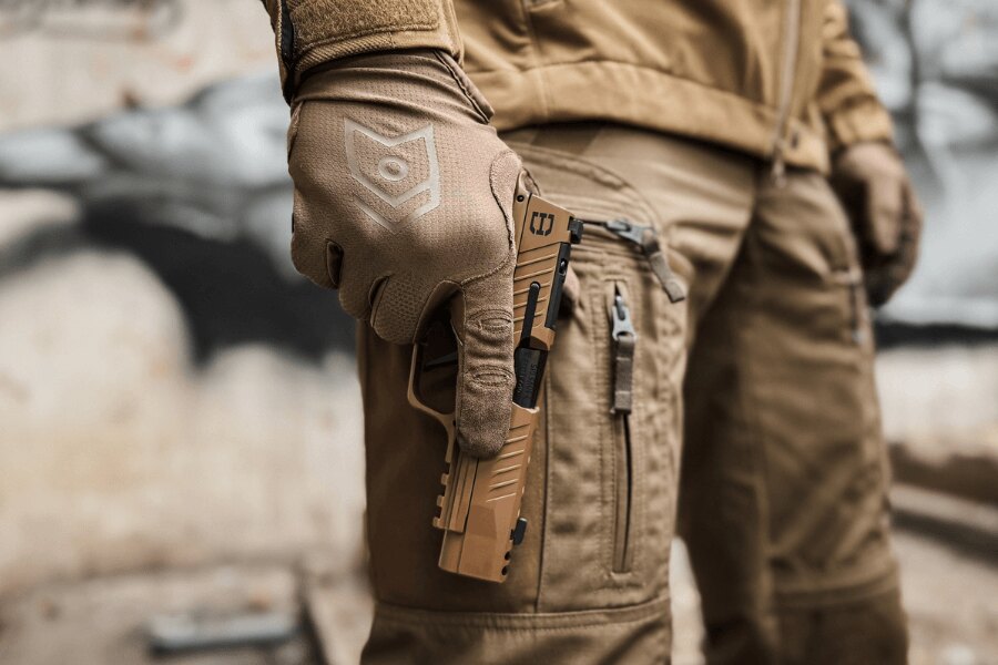 Střelec v pískové rukavici drží pistoli SIG Sauer v pískovém provedení podél stehna.