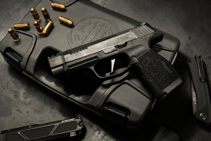 Černá pistole SIG Sauer P365 na přepravním kufru s logem výrobce, obklopená náboji, zavíracím nožem a svítilnou.