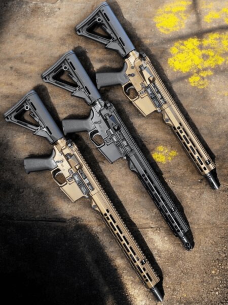 Detail černé pušky AR-15 s otevřenou krytkou výhozního okna mezi dvěma pískovými puškami.
