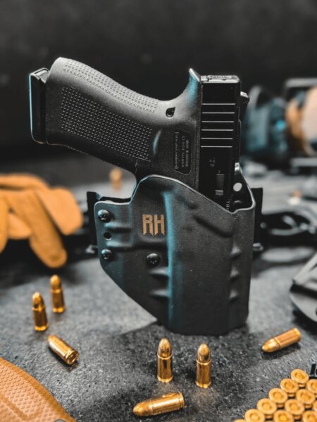Pistole Glock v černém vnějším pouzdře RH Holsters postavená na stole mezi náboji a střeleckým vybavením.