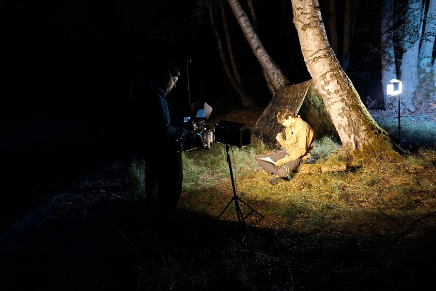 Noční natáčení Matěje Houšky před maskovací celtou v lese za světla filmových reflektorů.